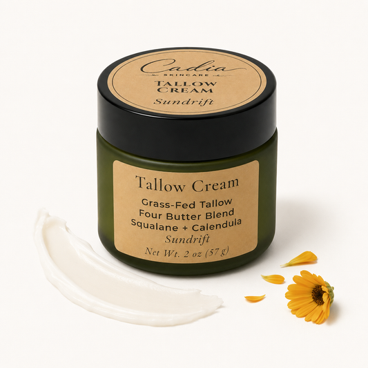 A jar of Cadia Skincare Sundrift Tallow Cream with a black lid and beige label, shown on a white background with a smear of white cream and yellow flower petals.